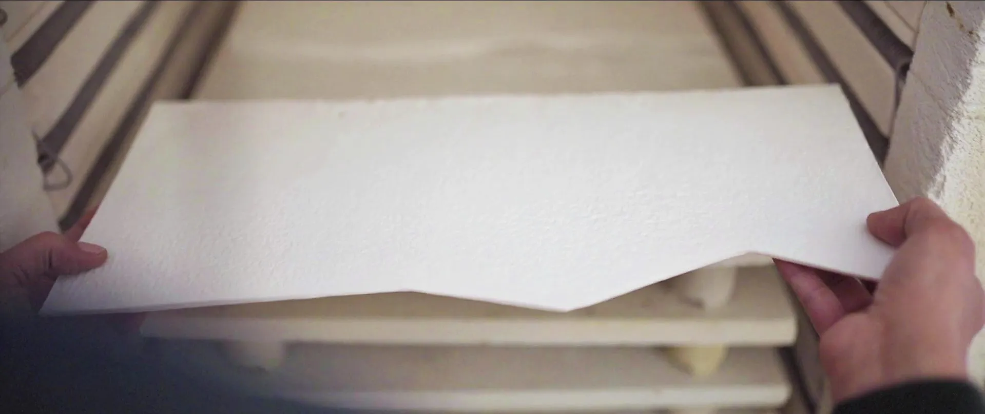 Deux mains sortent du four une plaque de porcelaine blanche, gauchie en son milieu comme un pli d’étoffe ; la surface garde le grain du façonnage à la main.