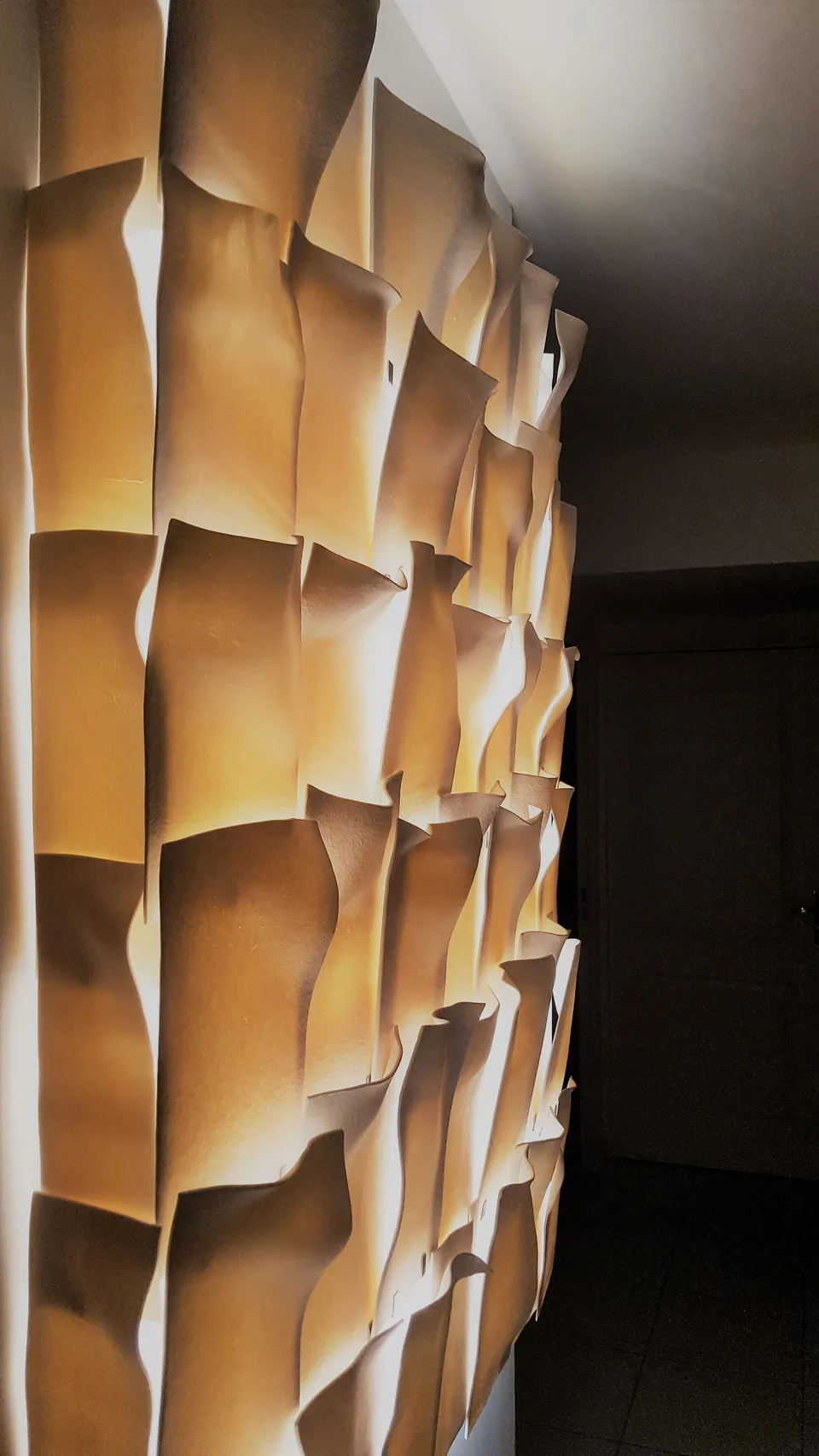 Mur de lumière vu de profil, composé de plaques de porcelaine décalées dont les plis passent du blanc à l’ambre puis à l’ombre.